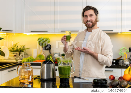 Middle-aged man headset showing fresh green smoothie from blender in home kitchen for healthy diet 140574994