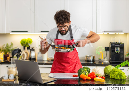Arabian young adult man watching laptop recipe while frying vegetables in home kitchen for meal prep 140575181