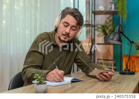 Middle-aged man listening audiobook headphones taking notes in notebook deep focused study at table 140575358
