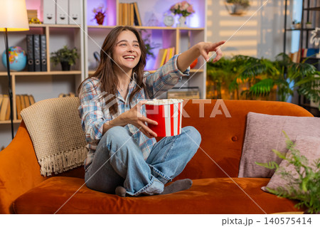 Young woman on home sofa eating popcorn while watching tv and laughing with cheerful playful mood Young woman on home sofa eating popcorn while watching tv and laughing with cheerful playful mood 140575414