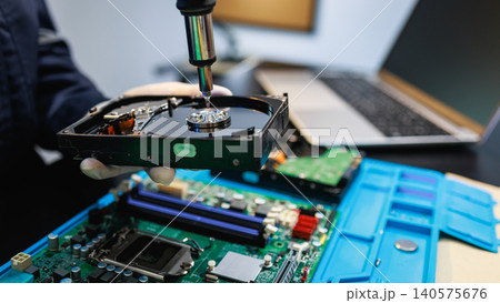 Close-up of a technician repairing a hard drive above a computer motherboard on a workbench, showing data recovery, hardware maintenance and electronics service. Close-up of a technician repairing a hard drive above a computer motherboard on a workbench, showing data recovery, hardware maintenance and electronics service. 140575676