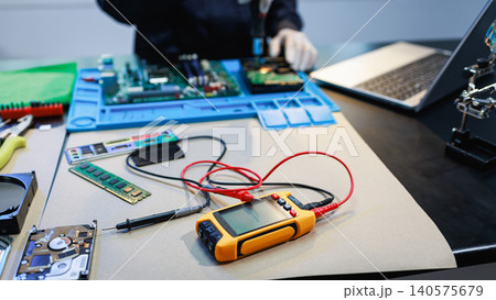 Digital multimeter and computer parts on a repair desk, with a technician working in the background. Ideal for electronics service, hardware testing, maintenance, and technology support. Digital multimeter and computer parts on a repair desk, with a technician working in the background. Ideal for electronics service, hardware testing, maintenance, and technology support. 140575679