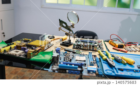 Computer repair workspace with circuit boards, motherboard parts, hand tools and a magnifying glass on a table in a bright room. Computer repair workspace with circuit boards, motherboard parts, hand tools and a magnifying glass on a table in a bright room. 140575683