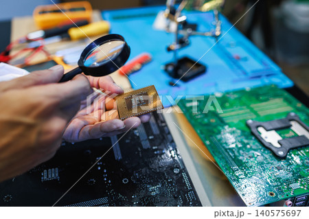 Close-up of hands inspecting a computer processor with a magnifying glass on a workbench with circuit boards and repair tools. 140575697