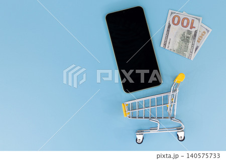 Minimalist scene with smartphone and shopping cart. Flat lay on solid blue background. Modern device beside miniature retail cart. Suggests online shopping or e-commerce concept 140575733