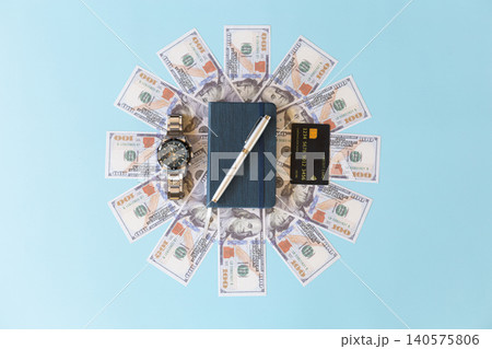 High angle view of a blue notebook and silver pen centered in a sunburst pattern of US dollar bills, accompanied by a luxury wristwatch and credit card on a blue background High angle view of a blue notebook and silver pen centered in a sunburst pattern of US dollar bills, accompanied by a luxury wristwatch and credit card on a blue background 140575806