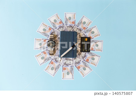 High angle view of a blue notebook and silver pen centered in a sunburst pattern of US dollar bills, accompanied by a luxury wristwatch and credit card on a blue background High angle view of a blue notebook and silver pen centered in a sunburst pattern of US dollar bills, accompanied by a luxury wristwatch and credit card on a blue background 140575807
