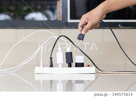 Hand plugs in charger into power strip. White surface reflects cables and devices. Multiple cords connect to outlets. No visible emotion or facial expression. Ideal for tech, home, or power management Hand plugs in charger into power strip. White surface reflects cables and devices. Multiple cords connect to outlets. No visible emotion or facial expression. Ideal for tech, home, or power management 140575814