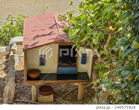 A small wooden cat house sits in a sunny garden. It has a red roof, white walls, and blue trim. Two pet bowls are placed nearby. Plants surround the house. A small wooden cat house sits in a sunny garden. It has a red roof, white walls, and blue trim. Two pet bowls are placed nearby. Plants surround the house. 140575928
