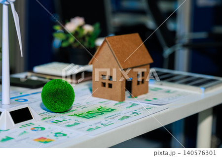 Small model house on energy charts beside a wind turbine and green plant ball on an office desk, showing eco home planning, renewable power, and sustainable real estate. Small model house on energy charts beside a wind turbine and green plant ball on an office desk, showing eco home planning, renewable power, and sustainable real estate. 140576301