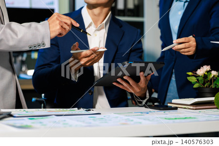 Business team in suits meeting at office desk, discussing notes and documents with tablet and pen during a planning session. Business team in suits meeting at office desk, discussing notes and documents with tablet and pen during a planning session. 140576303