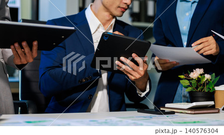 Business team in a modern office reviewing papers and tablets during a meeting, with hands visible, desk items and a small flower pot creating a professional workplace scene. Business team in a modern office reviewing papers and tablets during a meeting, with hands visible, desk items and a small flower pot creating a professional workplace scene. 140576304