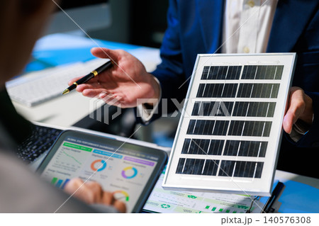 Business meeting about solar energy, with a professional showing a small solar panel beside a laptop with charts and reports in a modern office. Business meeting about solar energy, with a professional showing a small solar panel beside a laptop with charts and reports in a modern office. 140576308