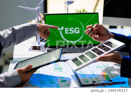 Business team discussing esg strategy at office desk with laptop screen, charts, tablet, solar panel sample and wind turbine models, showing sustainable planning and green investment. Business team discussing esg strategy at office desk with laptop screen, charts, tablet, solar panel sample and wind turbine models, showing sustainable planning and green investment. 140576317