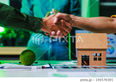 Close view of two people shaking hands over a desk with a small model house, showing a home sale, rental deal, mortgage approval, or real estate partnership in a modern office. 140576365