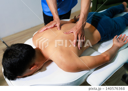Therapist giving a back massage to a young man lying on a treatment table in a wellness clinic, showing relaxation, body care, pain relief and professional therapy. 140576368