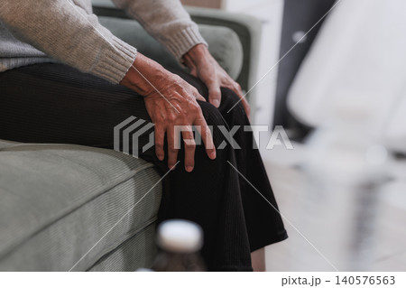 Close view of an older person sitting on a sofa with hands on knees, suggesting knee pain, joint stiffness, aging, health concerns, recovery, or home care. Close view of an older person sitting on a sofa with hands on knees, suggesting knee pain, joint stiffness, aging, health concerns, recovery, or home care. 140576563