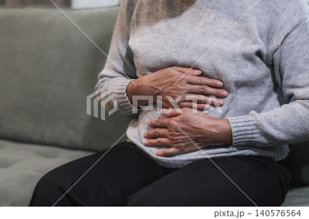 Close-up of an older woman sitting on a sofa and holding her stomach in pain at home, showing abdominal discomfort, cramps, digestion problems, or illness. Close-up of an older woman sitting on a sofa and holding her stomach in pain at home, showing abdominal discomfort, cramps, digestion problems, or illness. 140576564