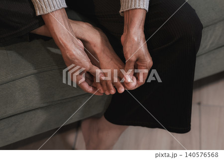 Close-up of a person sitting on a sofa and holding a painful foot at home, showing discomfort, cramp, injury or health problem in a calm indoor setting. Close-up of a person sitting on a sofa and holding a painful foot at home, showing discomfort, cramp, injury or health problem in a calm indoor setting. 140576568