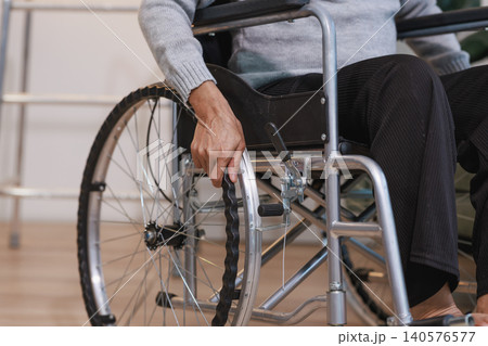 Close view of a senior person sitting in a wheelchair indoors, holding the wheel rim. Image suggests mobility support, recovery, disability care, independence and daily life at home or clinic. 140576577