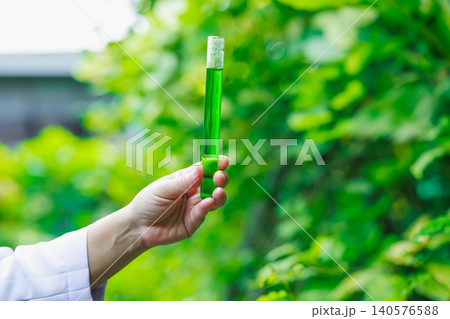 Hand holding a glass test tube filled with green liquid outdoors, with soft blurred leaves in the background, showing science, nature, research, and eco friendly ideas. Hand holding a glass test tube filled with green liquid outdoors, with soft blurred leaves in the background, showing science, nature, research, and eco friendly ideas. 140576588