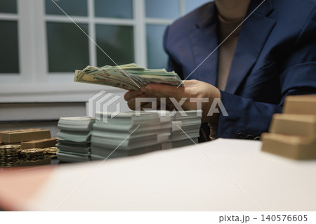 Close view of a person in a blue suit counting cash at a desk with stacks of banknotes and coins, showing wealth, finance, savings, business success, and money management. Close view of a person in a blue suit counting cash at a desk with stacks of banknotes and coins, showing wealth, finance, savings, business success, and money management. 140576605