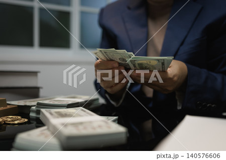 Person in a dark suit counting us dollar bills at a desk with stacks of cash and coins in an office, showing business, wealth, payment, savings, or financial planning. Person in a dark suit counting us dollar bills at a desk with stacks of cash and coins in an office, showing business, wealth, payment, savings, or financial planning. 140576606