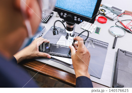 Technician repairs a smartphone on a workbench using a microscope and tools in an electronics service lab. Close view of device maintenance, testing and mobile phone fix. Technician repairs a smartphone on a workbench using a microscope and tools in an electronics service lab. Close view of device maintenance, testing and mobile phone fix. 140576625