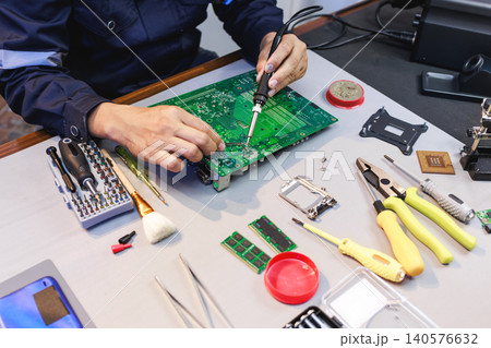 Technician repairing a green circuit board with a soldering iron at a workbench, surrounded by tools and computer parts in a clean electronics workspace. Technician repairing a green circuit board with a soldering iron at a workbench, surrounded by tools and computer parts in a clean electronics workspace. 140576632
