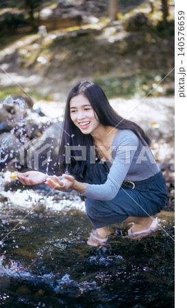 Smiling young woman crouching in a clear stream, splashing water with hands in a sunny forest setting; candid lifestyle outdoor nature scene. Smiling young woman crouching in a clear stream, splashing water with hands in a sunny forest setting; candid lifestyle outdoor nature scene. 140576659
