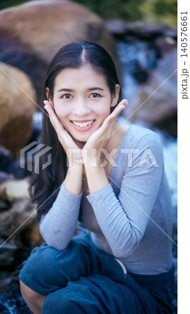 Smiling young woman posing outdoors by rocky stream, hands framing face, natural light, shallow depth of field, travel and lifestyle portrait. 140576661
