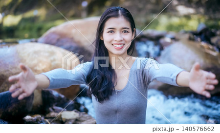 Smiling young woman with arms outstretched inviting a hug outdoors by rocky stream and waterfall, natural light, shallow depth of field, friendly welcoming gesture 140576662