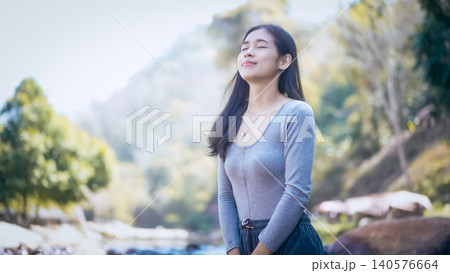 Young woman enjoying fresh air outdoors by a riverside, eyes closed, calm smile, soft morning light with blurred forest mountains in background 140576664