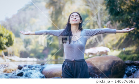 Young woman with arms outstretched enjoying fresh air by a river in a green forest, serene outdoor wellness moment in soft daylight. Young woman with arms outstretched enjoying fresh air by a river in a green forest, serene outdoor wellness moment in soft daylight. 140576665