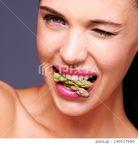 Woman face, lipstick and vegetables in mouth for beauty, flirting and vegan cosmetic product. Cruelty free makeup, portrait and wink, asparagus with pink lip gloss for aesthetic on purple background 140577680