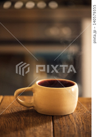 Ceramic coffee cup with steam rising, placed on a wooden table, blurred kitchen background with soft lighting creating a warm atmosphere Ceramic coffee cup with steam rising, placed on a wooden table, blurred kitchen background with soft lighting creating a warm atmosphere 140579355