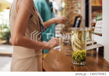 Woman in beige dress observes tea preparation with glass cup of green tea and tea leaves on wooden counter in a vibrant tea shop setting Woman in beige dress observes tea preparation with glass cup of green tea and tea leaves on wooden counter in a vibrant tea shop setting 140579361
