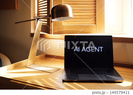 Laptop displaying text "AI AGENT" on a wooden desk with a modern lamp and window light illuminating the workspace in a contemporary office setting 140579411