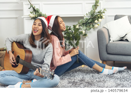 Happy Asian Woman sitting Christmas gift box under xmas tree playing acoustic guitar together. Two Women smile talk sitting decor christmas ball tree xmas holiday winter festival in living room Happy Asian Woman sitting Christmas gift box under xmas tree playing acoustic guitar together. Two Women smile talk sitting decor christmas ball tree xmas holiday winter festival in living room 140579634