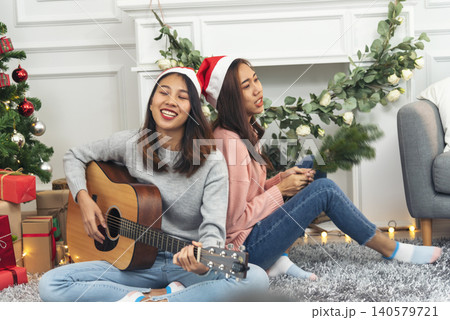 Happy Asian Woman sitting Christmas gift box under xmas tree playing acoustic guitar together. Two Women smile talk sitting decor christmas ball tree xmas holiday winter festival in living room Happy Asian Woman sitting Christmas gift box under xmas tree playing acoustic guitar together. Two Women smile talk sitting decor christmas ball tree xmas holiday winter festival in living room 140579721