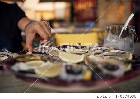 Hand reaching for fresh oysters on a plate garnished with lemon slices, with a glass of ice water and a blurred outdoor dining setting in the background Hand reaching for fresh oysters on a plate garnished with lemon slices, with a glass of ice water and a blurred outdoor dining setting in the background 140579939