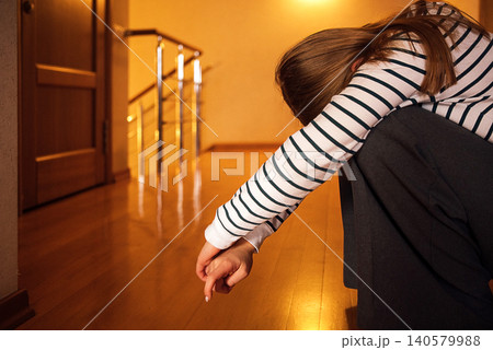 Young woman with long brown hair in striped shirt sits on wooden floor, leaning forward with arms resting on knees in a softly lit indoor setting Young woman with long brown hair in striped shirt sits on wooden floor, leaning forward with arms resting on knees in a softly lit indoor setting 140579988