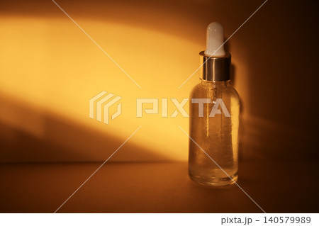 Clear glass dropper bottle with white pipette cap filled with liquid, illuminated by warm golden light against a smooth orange background Clear glass dropper bottle with white pipette cap filled with liquid, illuminated by warm golden light against a smooth orange background 140579989