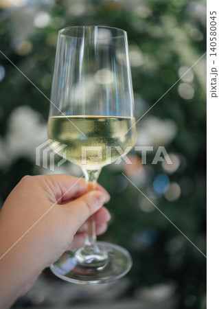 Hand holding a clear wine glass filled with white wine against a blurred background of greenery and decorative lights in a festive setting Hand holding a clear wine glass filled with white wine against a blurred background of greenery and decorative lights in a festive setting 140580045