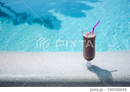 Chocolate smoothie in a tall glass with a pink straw placed on a stone ledge beside a clear blue swimming pool reflecting sunlight Chocolate smoothie in a tall glass with a pink straw placed on a stone ledge beside a clear blue swimming pool reflecting sunlight 140580048