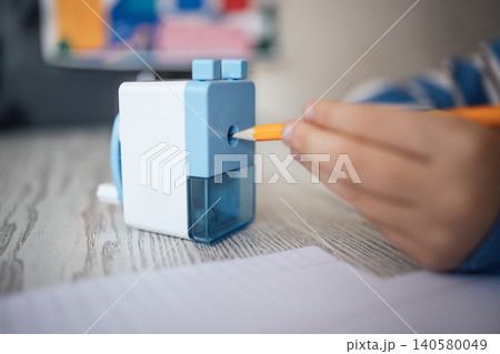 Child's hand holding orange pencil inserting it into a blue and white pencil sharpener on a wooden desk with blank paper and colorful artwork in the background Child's hand holding orange pencil inserting it into a blue and white pencil sharpener on a wooden desk with blank paper and colorful artwork in the background 140580049