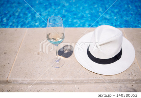 Glass of white wine and a white sun hat with black band resting on stone edge beside a clear blue swimming pool under bright sunlight Glass of white wine and a white sun hat with black band resting on stone edge beside a clear blue swimming pool under bright sunlight 140580052