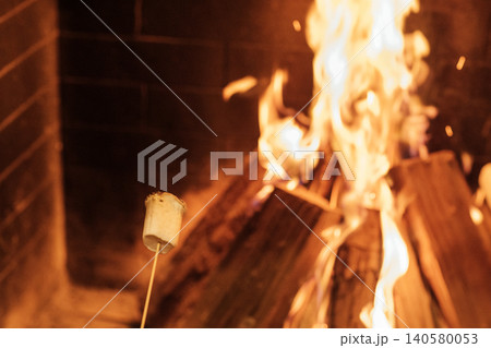 Marshmallow on a stick roasting over an open flame in a fireplace, with logs burning brightly and glowing embers visible in the background 140580053