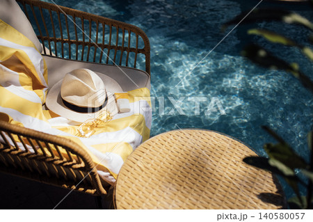Woven rattan chair with yellow striped towel, sun hat, and sunglasses beside a sparkling blue swimming pool in a tropical outdoor setting Woven rattan chair with yellow striped towel, sun hat, and sunglasses beside a sparkling blue swimming pool in a tropical outdoor setting 140580057