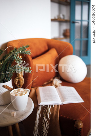 Cozy living room scene featuring an orange velvet sofa, a warm blanket, a cup of hot chocolate with marshmallows, and an open book on a marble table, creating a relaxing atmosphere Cozy living room scene featuring an orange velvet sofa, a warm blanket, a cup of hot chocolate with marshmallows, and an open book on a marble table, creating a relaxing atmosphere 140580074